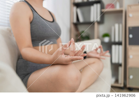 Close-up of a woman practicing meditation in a cozy living room, focusing on her hands in a mudra gesture while seated on a sofa 141962906