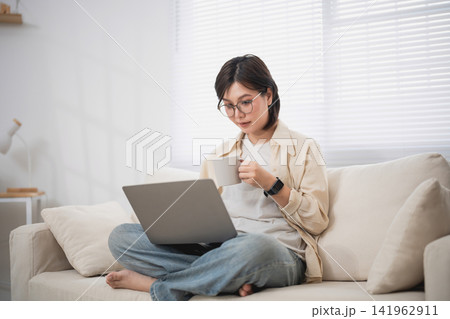 Woman Relaxing on Couch with Laptop and Drinking Coffee in Bright Living Room Woman Relaxing on Couch with Laptop and Drinking Coffee in Bright Living Room 141962911