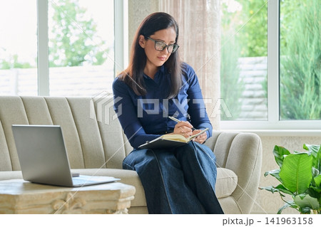 Attentive businesswoman taking notes in notebook while looking at laptop screen at home office Attentive businesswoman taking notes in notebook while looking at laptop screen at home office 141963158