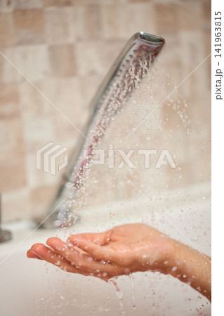 Person's hand feeling refreshing water droplets pouring from a shower head in a bathroom, symbolizing hygiene and cleanliness Person's hand feeling refreshing water droplets pouring from a shower head in a bathroom, symbolizing hygiene and cleanliness 141963815