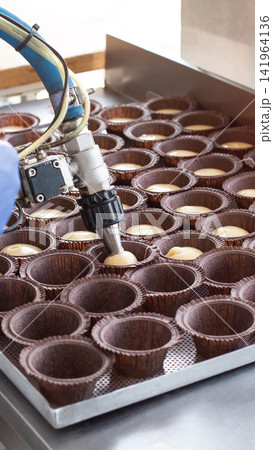 Industrial robotic arm dispensing cupcake batter into paper molds on metal baking tray, automated dessert production line, bakery manufacturing process vertical photo Industrial robotic arm dispensing cupcake batter into paper molds on metal baking tray, automated dessert production line, bakery manufacturing process vertical photo 141964136