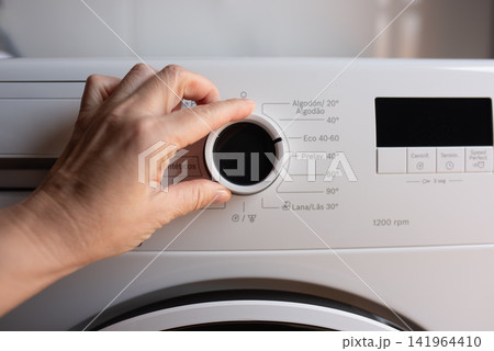 A hand adjusts the circular program selection knob on a white washing machine with Russian text. A hand adjusts the circular program selection knob on a white washing machine with Russian text. 141964410