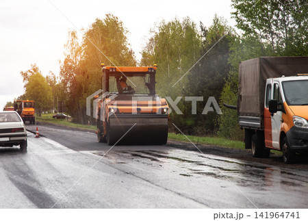 Road roller compacting fresh asphalt layer during construction work on suburban highway. Cargo truck parked at roadside, cars driving past. Copy space for text Road roller compacting fresh asphalt layer during construction work on suburban highway. Cargo truck parked at roadside, cars driving past. Copy space for text 141964741