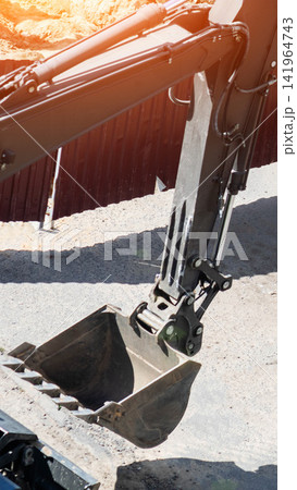 Excavator arm and bucket at construction site on sunny day, heavy machinery preparing ground for building development or infrastructure project with bright sun flare. Vertical photo Excavator arm and bucket at construction site on sunny day, heavy machinery preparing ground for building development or infrastructure project with bright sun flare. Vertical photo 141964743