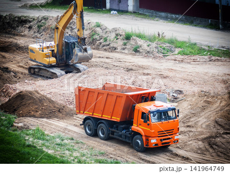 Orange dump truck and yellow excavator on construction site, heavy machinery working on earthmoving project, industrial equipment for land development and building Orange dump truck and yellow excavator on construction site, heavy machinery working on earthmoving project, industrial equipment for land development and building 141964749