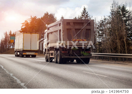 Heavy duty dump truck and white semi trailer truck driving on asphalt highway road with evergreen and deciduous trees under cloudy sky, 40-ton truck 141964750
