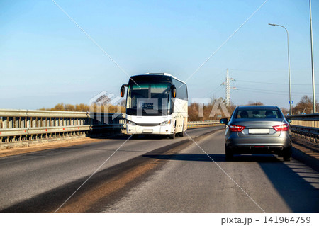 White tour bus traveling on bridge roadway towards camera perspective alongside grey car from rear. Clear blue sky and bare trees frame background scene. White tour bus traveling on bridge roadway towards camera perspective alongside grey car from rear. Clear blue sky and bare trees frame background scene. 141964759