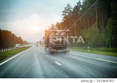 Heavy transport truck carrying new vehicles on multi-lane highway during rain, wet road surface, spray from tires, reduced visibility of road ahead. Copy space for text, custom-made car delivery Heavy transport truck carrying new vehicles on multi-lane highway during rain, wet road surface, spray from tires, reduced visibility of road ahead. Copy space for text, custom-made car delivery 141964765