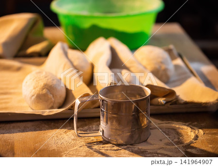 Unbaked dough balls and pastry sheets on wooden surface with flour sifter, ready for baking preparation in rustic kitchen setting, close up of culinary ingredients Unbaked dough balls and pastry sheets on wooden surface with flour sifter, ready for baking preparation in rustic kitchen setting, close up of culinary ingredients 141964907
