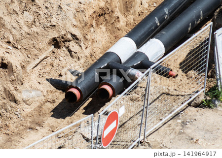 Black heating mains pipes with insulation exposed in a construction trench during repair and relocation work, with temporary fencing and a no entry sign protecting site. Black heating mains pipes with insulation exposed in a construction trench during repair and relocation work, with temporary fencing and a no entry sign protecting site. 141964917