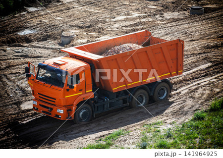 Orange heavy duty dump truck on construction site filled with rubble, ready for transport, ground uneven with tire tracks, bright sunny day, outdoor industrial scene. Orange heavy duty dump truck on construction site filled with rubble, ready for transport, ground uneven with tire tracks, bright sunny day, outdoor industrial scene. 141964925
