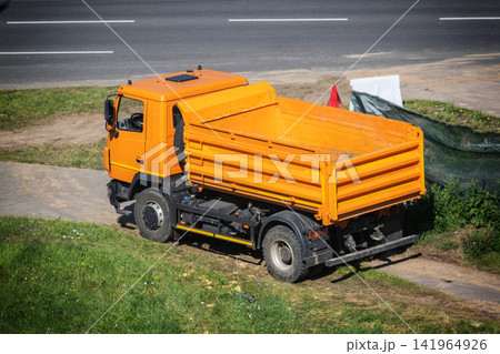 Orange tipper truck parked on dirt path next to asphalt road with white lines, vehicle ready for construction work or material transport, bright sunny day, overhead view of utility truck. Orange tipper truck parked on dirt path next to asphalt road with white lines, vehicle ready for construction work or material transport, bright sunny day, overhead view of utility truck. 141964926