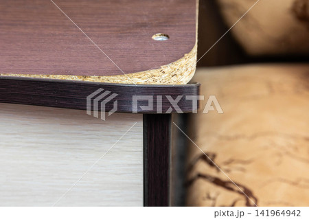 Close-up detail of edge of particle board furniture showing exposed chipboard core and peeling laminate surface, screw head visible, worn corner construction of cabinet lid or shelf Close-up detail of edge of particle board furniture showing exposed chipboard core and peeling laminate surface, screw head visible, worn corner construction of cabinet lid or shelf 141964942