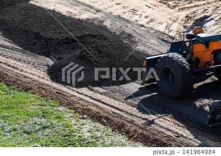 Heavy construction machinery grader levels surface of new road, spreading and compacting fresh black asphalt chips for infrastructure development work on sunny day. 141964984