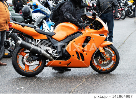 Orange sport motorcycle parked on wet asphalt, rider in black gear and helmet stands beside vehicle, blurred background shows many other bikes and people gathered at outdoor event. Orange sport motorcycle parked on wet asphalt, rider in black gear and helmet stands beside vehicle, blurred background shows many other bikes and people gathered at outdoor event. 141964997