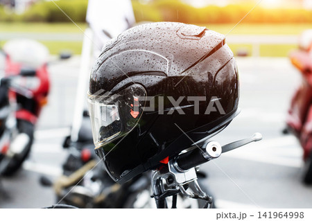 Black motorcycle helmet resting on handlebars of bike outdoors after rain, covered in water droplets with blurry background and sunlight flare, safety equipment for rider. Black motorcycle helmet resting on handlebars of bike outdoors after rain, covered in water droplets with blurry background and sunlight flare, safety equipment for rider. 141964998