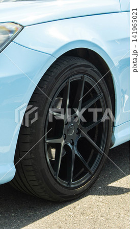 Light blue luxury sport car front fender and glossy black multi-spoke custom alloy wheel with low profile performance tire. Vehicle detail on asphalt street during sunny day, vertical composition Light blue luxury sport car front fender and glossy black multi-spoke custom alloy wheel with low profile performance tire. Vehicle detail on asphalt street during sunny day, vertical composition 141965021