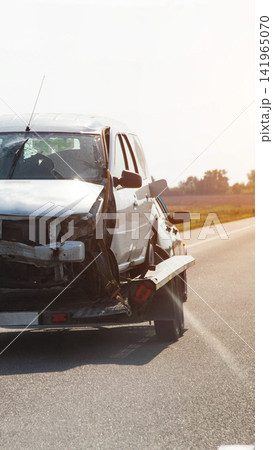 Damaged white vehicle loaded on tow truck after accident, broken windshield and crushed front end visible on highway road, sunny day vertical photo Damaged white vehicle loaded on tow truck after accident, broken windshield and crushed front end visible on highway road, sunny day vertical photo 141965070