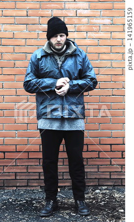 Adult Caucasian man full body portrait standing against brick wall, wearing winter clothing including beanie and jacket, clasping hands, somber expression, outdoor scene, vertical photo 141965159