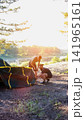 Two adult men assembling camping tent by serene lake water with lush green trees at golden hour sunset light, preparing for outdoor adventure, vertical photo 141965161