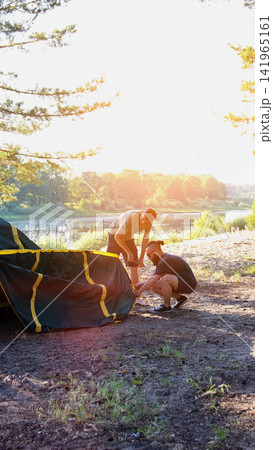 Two adult men assembling camping tent by serene lake water with lush green trees at golden hour sunset light, preparing for outdoor adventure, vertical photo 141965161