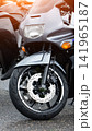 Front view of parked sport motorcycle with grey and black body, showing wheel, brake disc, and suspension after rain, drops visible, sunlight reflection on upper part. 141965187