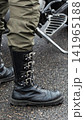 Close up view of person leg with heavy black combat boot featuring multiple silver spikes and buckles, olive green cargo pants, wet asphalt surface, motorcycle footpeg visible, vertical photo format 141965188