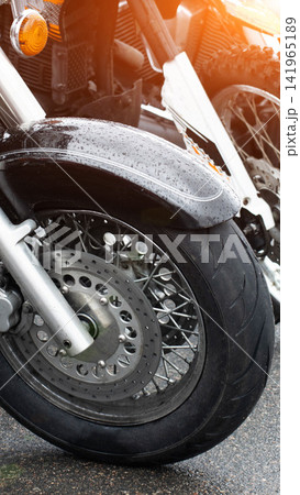 Motorcycle front wheel, fender, and disc brake detail with raindrops on glossy surface under warm sunlight flare, vertical shot of vehicle parts Motorcycle front wheel, fender, and disc brake detail with raindrops on glossy surface under warm sunlight flare, vertical shot of vehicle parts 141965189
