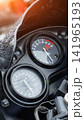 Close-up detail of motorcycle dashboard with speedometer showing speed in kilometers per hour and tachometer displaying engine RPM, raindrops visible on windshield, bright lens flare top left 141965193