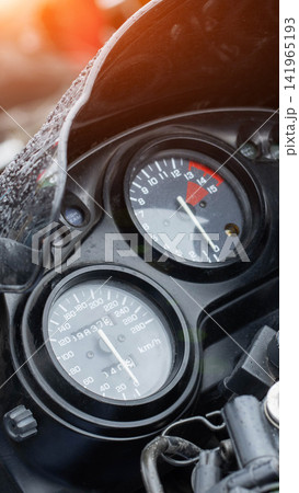 Close-up detail of motorcycle dashboard with speedometer showing speed in kilometers per hour and tachometer displaying engine RPM, raindrops visible on windshield, bright lens flare top left Close-up detail of motorcycle dashboard with speedometer showing speed in kilometers per hour and tachometer displaying engine RPM, raindrops visible on windshield, bright lens flare top left 141965193