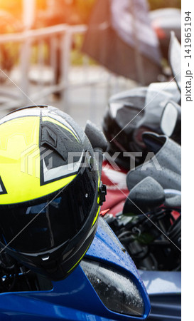 Neon yellow and black motorcycle helmet with raindrops on surface of blue motorbike parked outdoors. Sunlight shining on helmet and wet vehicle, reflecting light from wet surfaces. Ready for ride. 141965194