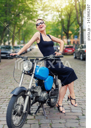 Portrait of pretty young adult stylish elegant woman wear black dress sit vintage retro blue motorcycle moped european city street Germany. Happy female person model enjoy having fun urban outside Portrait of pretty young adult stylish elegant woman wear black dress sit vintage retro blue motorcycle moped european city street Germany. Happy female person model enjoy having fun urban outside 141966297