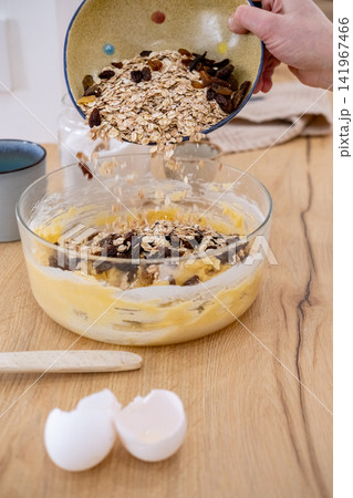 Homemade breakfast process. Healthy recipe. Female chef hand pouring oat flakes raisins in dough bowl on kitchen counter with eggshell. Homemade breakfast process. Healthy recipe. Female chef hand pouring oat flakes raisins in dough bowl on kitchen counter with eggshell. 141967466