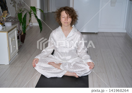 Woman Sitting Cross Legged On Black Exercise Mat With Hands Resting On Knees Indoors, Practicing Woman Sitting Cross Legged On Black Exercise Mat With Hands Resting On Knees Indoors, Practicing 141968264