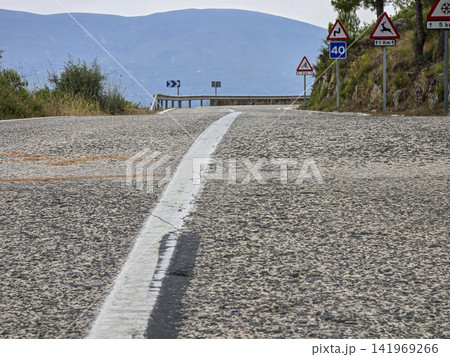 Low angle view of a narrow mountain pass road with sharp turn signs and steep cliffs ahead in a rural area Low angle view of a narrow mountain pass road with sharp turn signs and steep cliffs ahead in a rural area 141969266