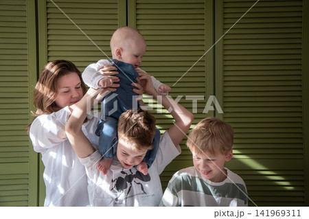 Mother helps baby balance on older brothers shoulders while three sons play together at home. Motherhood, brotherhood, sibling care, parenting, childhood, family bond, support, play, togetherness Mother helps baby balance on older brothers shoulders while three sons play together at home. Motherhood, brotherhood, sibling care, parenting, childhood, family bond, support, play, togetherness 141969311