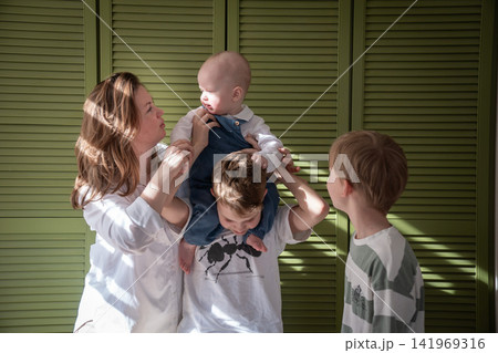 Mother poses with three sons while baby sits on older brothers shoulders during playful family moment at home. Motherhood, brotherhood, sibling bond, parenting, childhood, family connection Mother poses with three sons while baby sits on older brothers shoulders during playful family moment at home. Motherhood, brotherhood, sibling bond, parenting, childhood, family connection 141969316