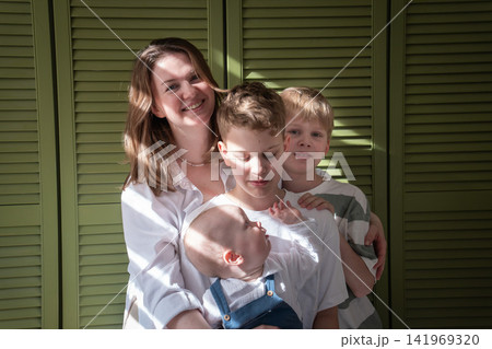 Mother embraces three sons while baby sits on older brothers lap during close family moment at home. Motherhood, brotherhood, sibling bond, parenting, childhood, family love, caregiving, togetherness Mother embraces three sons while baby sits on older brothers lap during close family moment at home. Motherhood, brotherhood, sibling bond, parenting, childhood, family love, caregiving, togetherness 141969320