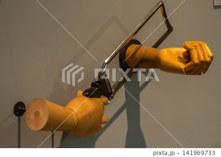 Wooden mannequin arms with hacksaw creating surreal self cutting wall installation Wooden mannequin arms with hacksaw creating surreal self cutting wall installation 141969733