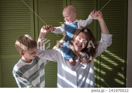 Mother lifts baby son on her shoulders and holds his hands while older brother plays beside them at home. Motherhood, parenting, childhood, family bonding, play, joy, affection, togetherness Mother lifts baby son on her shoulders and holds his hands while older brother plays beside them at home. Motherhood, parenting, childhood, family bonding, play, joy, affection, togetherness 141970022