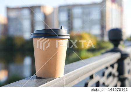 Kraft paper coffee cup mockup on bridge handrail with blurred city buildings and autumn trees in background. Perfect for coffee to go, urban autumn lifestyle, outdoor takeaway branding Kraft paper coffee cup mockup on bridge handrail with blurred city buildings and autumn trees in background. Perfect for coffee to go, urban autumn lifestyle, outdoor takeaway branding 141970161