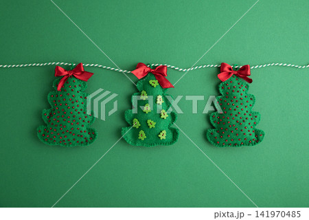 Three Festive Felt Christmas Tree Ornaments Hanging on White String Against Green Background Decorations 141970485