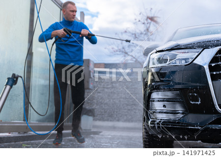Man cleans his vehicle at self service car wash station using professional washing equipment. Rinsing the vehicle with high-pressure water stream system. Outdoor washing bay Man cleans his vehicle at self service car wash station using professional washing equipment. Rinsing the vehicle with high-pressure water stream system. Outdoor washing bay 141971425