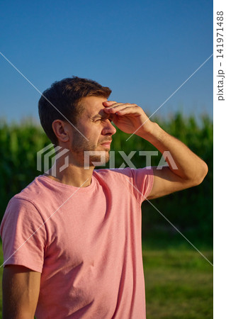 Man raises his hand over his eyes to block bright sunlight while standing near cornfield Man raises his hand over his eyes to block bright sunlight while standing near cornfield 141971488