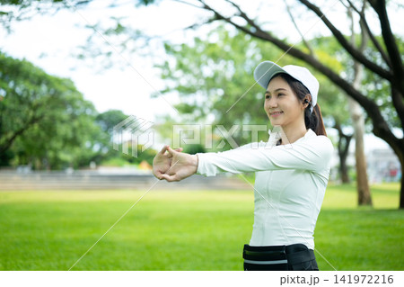 Young Asian Woman Stretching Arms in Green Park Before Workout, Smiling and Happy 141972216