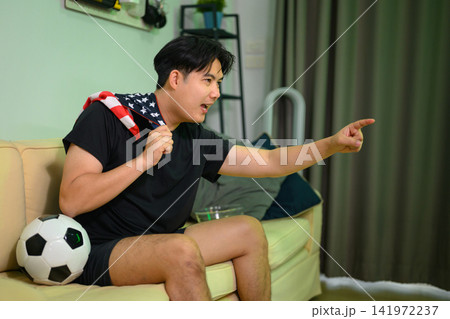 Excited Man Cheering Football Game at Home with American Flag and Soccer Ball on Sofa Excited Man Cheering Football Game at Home with American Flag and Soccer Ball on Sofa 141972237
