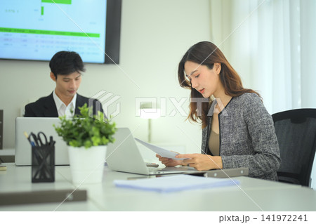Female professional handing reports to colleague in office meeting with presentation screen in background Female professional handing reports to colleague in office meeting with presentation screen in background 141972241