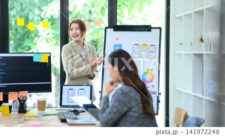 Young Gen Z Asian Female Developer Presenting Mobile App UI UX Design to Colleague on Whiteboard in Modern Office 141972248