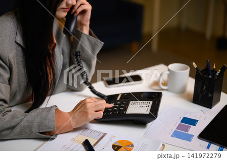 Businesswoman Talking on Landline Phone at Office Desk with Financial Charts and Documents 141972279