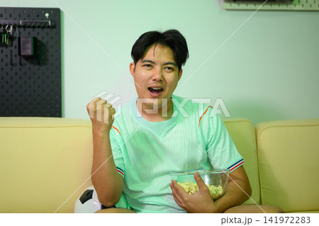 Excited Man Cheering Football Game at Home Holding Popcorn Bowl on Sofa with Soccer Ball Excited Man Cheering Football Game at Home Holding Popcorn Bowl on Sofa with Soccer Ball 141972283